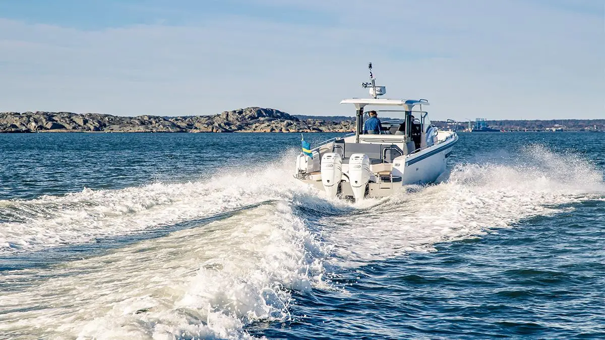 A motorboat with two outboard engines creates a wake as it navigates through calm water, with rocky coastline in the background.